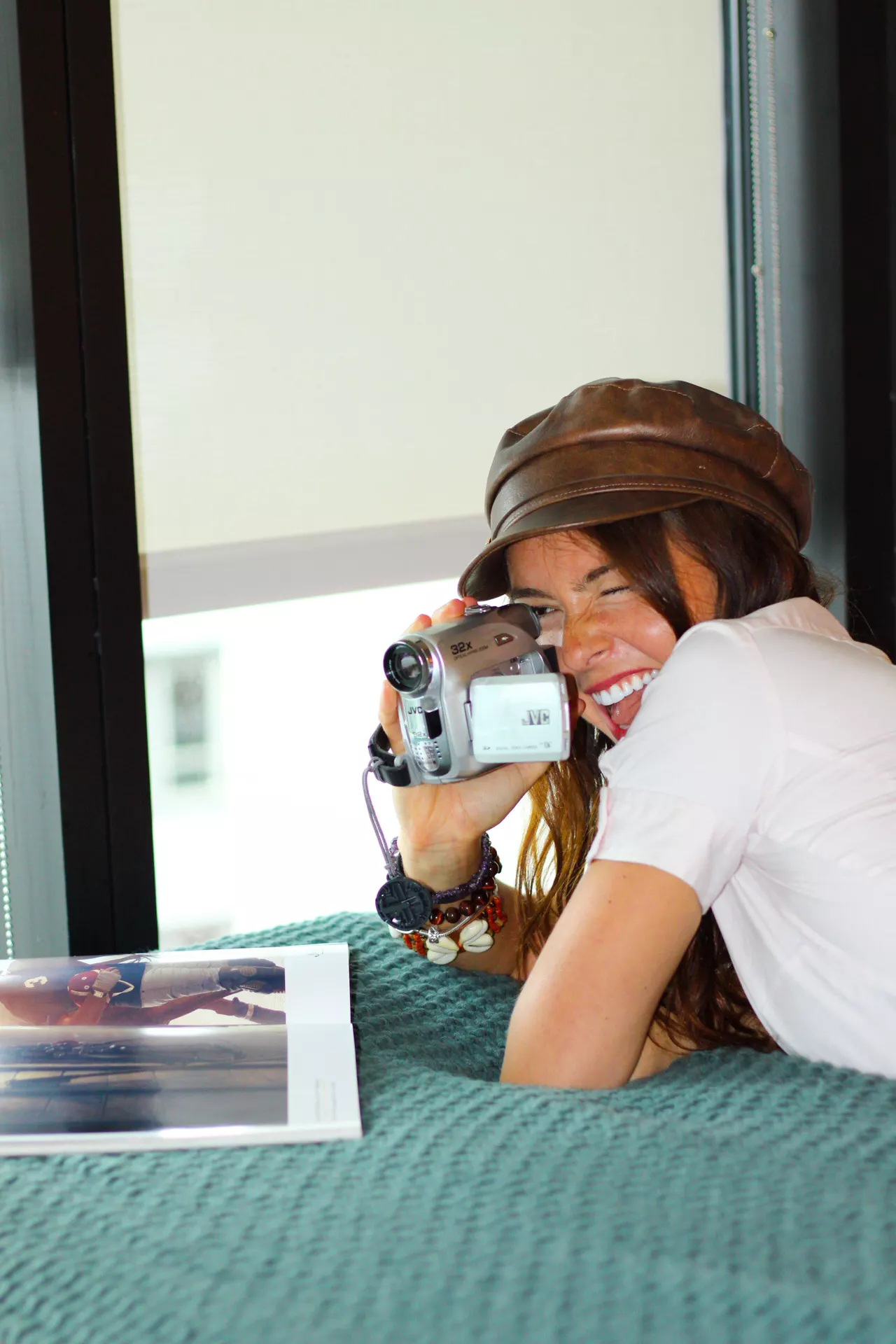 Smiling resident in a leather newsboy cap filming with a handheld camcorder while lying on a bed