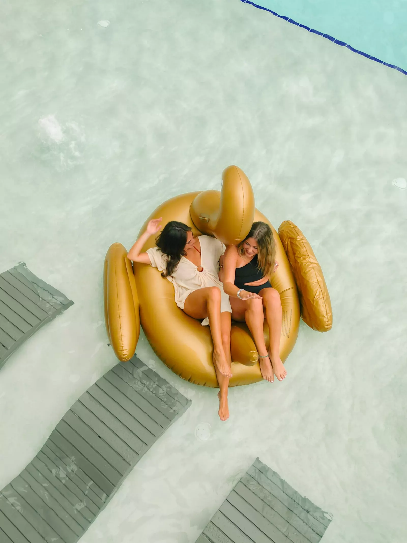 Overhead view of two friends laughing together on a gold swan float in the pool